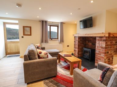 A living room with grey sofas a wooden coffee table a brick fireplace and a wall-mounted TV at Llety Dewi in Lake Vyrnwy