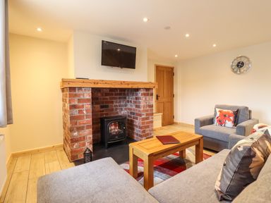 A living room with a brick fireplace and wood stove a wall-mounted TV grey sofas a wooden coffee table and a clock on the wall at Llety Dewi in Lake Vyrnwy