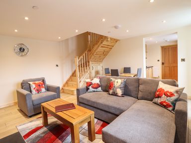 A living room with a gray sofa and armchair with floral cushions a wooden coffee table a patterned rug a wooden staircase and a dining area at Llety Dewi in Lake Vyrnwy