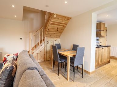 A dining area with a wooden table and four grey chairs next to wooden stairs and an open kitchen at Llety Dewi in Lake Vyrnwy