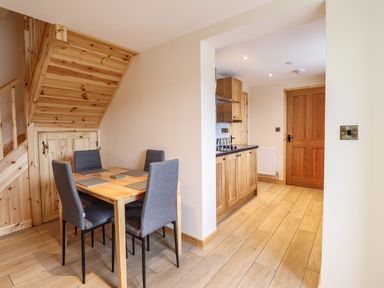 A dining area with a wooden table and four grey chairs next to wooden stairs and an entrance to a kitchen at Llety Dewi in Lake Vyrnwy
