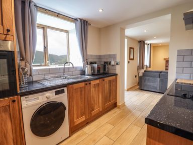 A kitchen with wooden cabinets a washing machine a sink and a microwave next to a living room with a sofa at Llety Dewi in Lake Vyrnwy