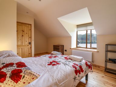 A bedroom with a bed covered in floral bedding two rolled towels on the bed a wooden door a wooden chest of drawers a large window with a view and a shelving unit at Llety Dewi in Lake Vyrnwy