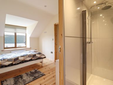 A bedroom with a bed next to a window overlooking hills and a lake and an adjacent shower at Llety Dewi in Lake Vyrnwy