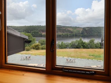 A view through a wooden window showing a gravel area with two tables and chairs overlooking a grassy field and a lake with forested hills at Old Cart House in Llanwddyn