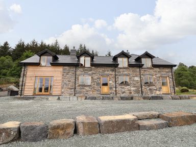 A stone house with four dormer windows and wooden doors surrounded by gravel and trees at Old Cart House in Llanwddyn