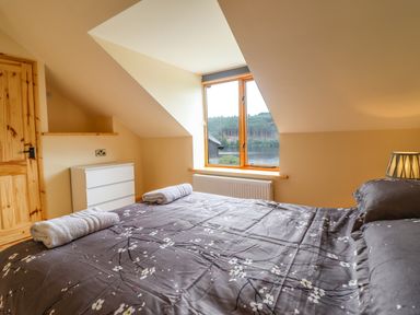 A bedroom with a bed covered in a floral patterned duvet two rolled towels on the bed a white dresser a wooden door and a window with a view of trees and water at Old Cart House in Llanwddyn
