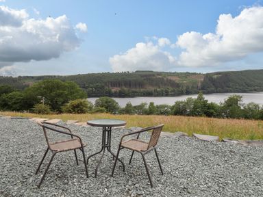 An outdoor seating area with two chairs and a table on gravel overlooking a lake and forested hills at Old Cart House in Llanwddyn