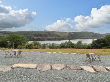 An outdoor seating area with tables and chairs overlooking a lake and forested hills at Old Cart House in Llanwddyn