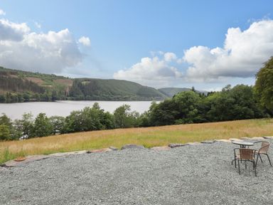 An outdoor area with a gravel surface, two chairs and a small table overlooking a lake and wooded hills at Old Cart House in Llanwddyn
