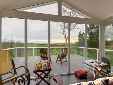 A sunroom with wooden chairs a small table and a blanket-covered bench overlooking a deck and green fields at The Schooner in Goleen County Cork