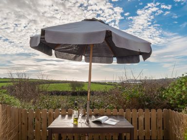 An outdoor wooden table with a bottle, two wine glasses, and a book under an umbrella with a view of fields and sky at The Schooner in Goleen County Cork
