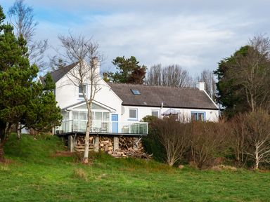A white house with a glass enclosed porch and stacked firewood under the deck surrounded by trees and grass at The Schooner in Goleen County Cork