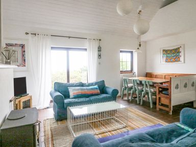 A living room with two blue sofas a glass coffee table a wood burning stove and a dining area with wooden bench and chairs at The Schooner in Goleen County Cork