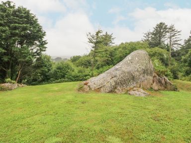 A grassy area with a large rock formation and trees in the background at The Schooner in Goleen County Cork
