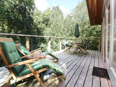 A wooden deck with green cushioned chairs a table with umbrella and surrounded by trees at 6 Streamside in Lanreath