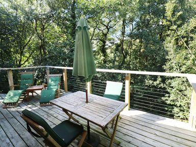 An outdoor wooden deck with green cushioned chairs a table and a closed green umbrella surrounded by trees at 6 Streamside in Lanreath