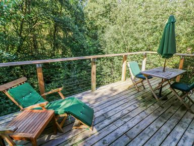 An outdoor wooden deck with reclining and regular chairs a table with an umbrella surrounded by trees at 6 Streamside in Lanreath