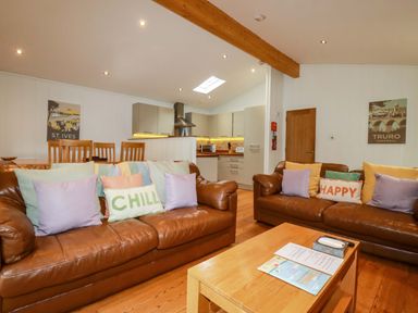 A living room with brown leather sofas and colorful cushions next to a wooden dining table and an open kitchen at 6 Streamside in Lanreath