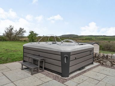 An outdoor hot tub with water jets on a stone patio with green fields and hills in the background at Hen Hafod in Talwrn near Llangefni