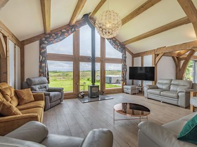 A living room with multiple sofas a wood-burning stove and large windows overlooking a green field at Hen Hafod in Talwrn near Llangefni