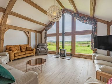A living room with large windows showing a green landscape a wood burning stove sofas armchairs a coffee table and exposed wooden beams at Hen Hafod in Talwrn near Llangefni