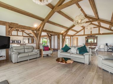 An open living room with wooden beams a gray leather sofa two armchairs a round copper coffee table and a dining table at Hen Hafod in Talwrn near Llangefni