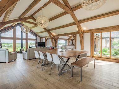 A dining area with a wooden table and white chairs next to a sitting area with sofas and large windows at Hen Hafod in Talwrn near Llangefni