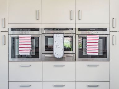 A kitchen wall with three built in stainless steel ovens and cabinets with towels hanging on oven handles at Hen Hafod in Talwrn near Llangefni