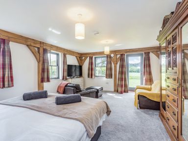 A bedroom with a bed with towels a leather chair an ottoman a television an armchair with a yellow cover wooden beams and curtains at Hen Hafod in Talwrn near Llangefni