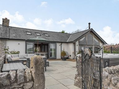 A patio with outdoor seating umbrella and hot tub in front of a building with stone wall at Hen Hafod in Talwrn near Llangefni