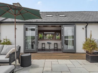 An outdoor patio with seating and an umbrella in front of open glass doors showing a kitchen bar at Hen Hafod in Talwrn near Llangefni
