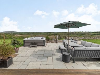 An outdoor patio with a hot tub a seating area with cushions a table an umbrella and potted plants at Hen Hafod in Talwrn near Llangefni