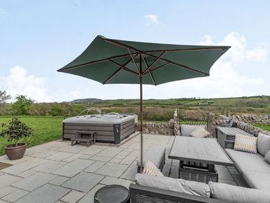A patio area with a hot tub a seating area with cushions and an umbrella at Hen Hafod Talwrn near Llangefni