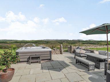 An outdoor patio with a hot tub table seating and an umbrella overlooking countryside at Hen Hafod in Talwrn near Llangefni