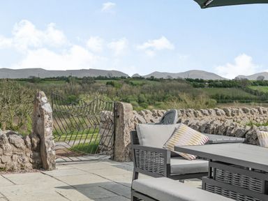 An outdoor patio with a table and chairs next to a stone wall and a metal gate overlooking green fields at Hen Hafod in Talwrn near Llangefni