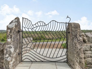 An iron gate with a leaf pattern between two stone pillars and a rural landscape in the background at Hen Hafod in Talwrn near Llangefni