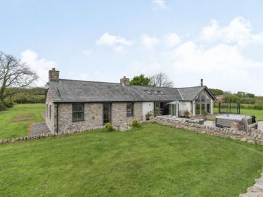 A stone house with multiple chimneys and a patio area with outdoor furniture and a hot tub at Hen Hafod in Talwrn near Llangefni