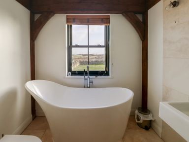 A freestanding bathtub under a window with wooden beams in a bathroom at Hen Hafod in Talwrn near Llangefni