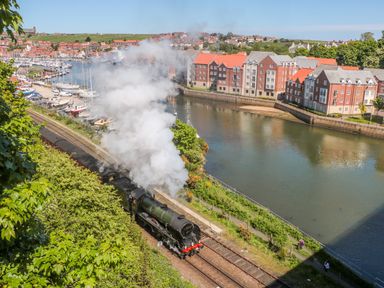 A steam train traveling along tracks next to a river with boats and buildings in the background at The Tannery in Whitby