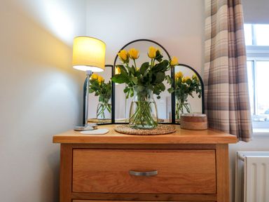 A wooden dresser with a three-panel mirror a vase of yellow flowers a table lamp and a window with plaid curtains at The Tannery in Whitby