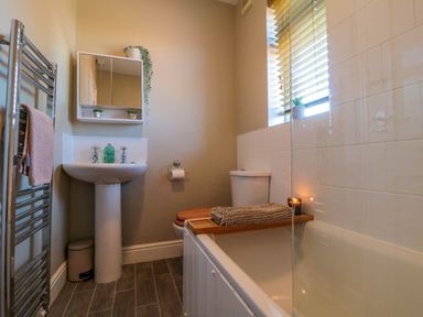 A bathroom with a pedestal sink a toilet a bathtub with a wooden tray a towel rack and a window at The Tannery in Whitby