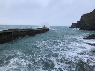 A stone pier extending into the ocean with waves crashing against it and rocky cliffs on the right at The Chace in Portreath