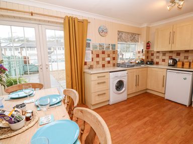 A kitchen with wooden cabinets a washing machine a fridge a dining table set with blue plates and glasses and yellow curtains at The Chace in Portreath