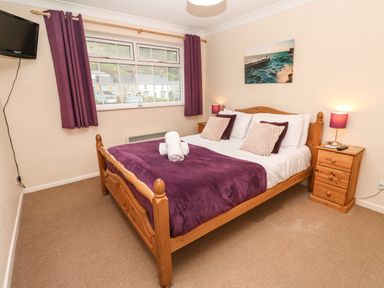 A bedroom with a wooden bed, purple bedding, purple curtains, two bedside tables with lamps, a wall-mounted TV, and a window showing houses outside at The Chace in Portreath