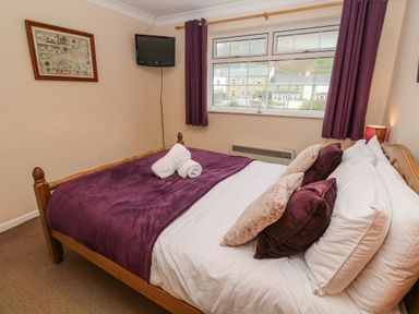A bedroom with a wooden bed with white sheets purple blankets and pillows a wall-mounted television and a large window with purple curtains at The Chace in Portreath