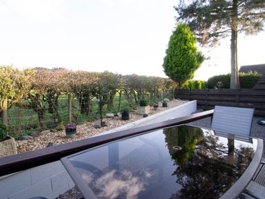 An outdoor patio area with a glass table and chairs overlooking a garden with trees and a wooden fence at Kestrel Lodge in Dumfries