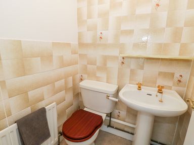 A bathroom with a toilet with wooden seat a pedestal sink with gold taps a radiator with a towel and tiled walls at Rose Cottage in Bream