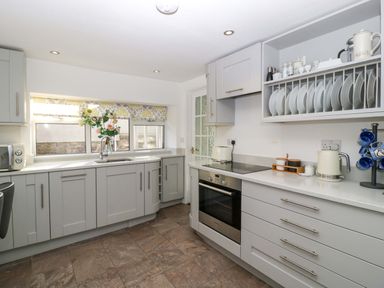 A kitchen with grey cabinets a built in oven a stove and a window with flowers on the sink at Rose Cottage in Bream