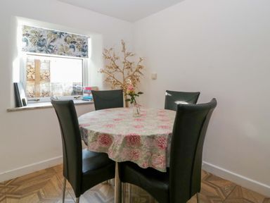 A dining area with a round table covered with a floral tablecloth and four black chairs by a window with a floral blind at Rose Cottage in Bream